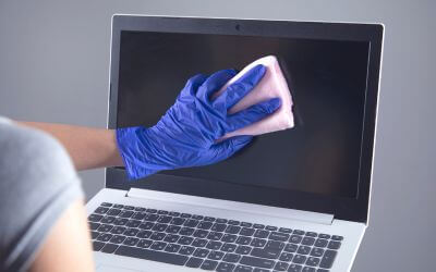 Laptop Monitor wird mit einem rosa Tuch gereinigt dass von einer Hand mit Putzhandschuhen gehalten wird